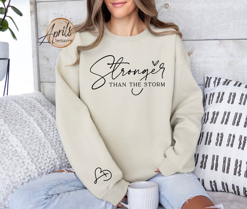 May include: A beige sweatshirt with the text "Stronger than the storm" and a small heart design. The sweatshirt has a small heart with a cross inside on the sleeve.