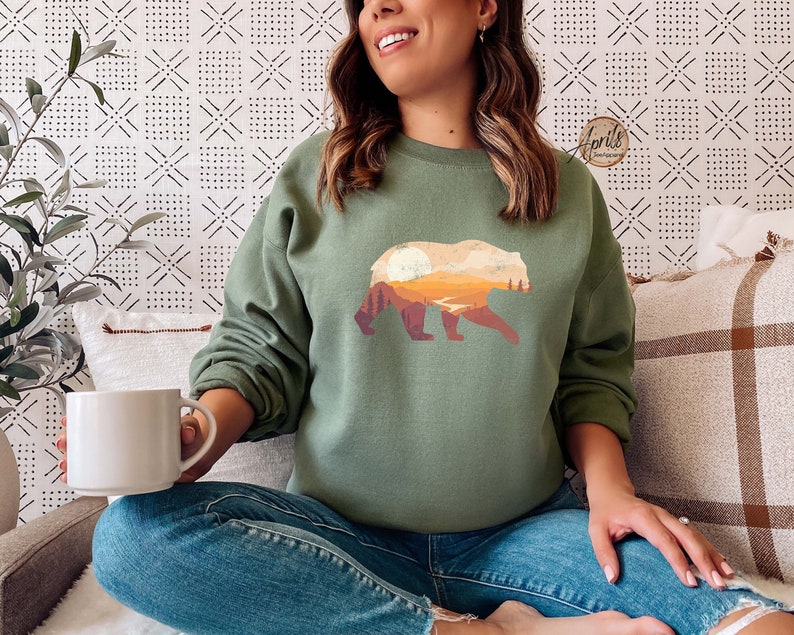 May include: A green sweatshirt with a graphic print of a bear standing in front of a mountain range with a sunset in the background.