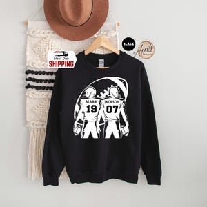 May include: Black sweatshirt featuring a white graphic of two football players with helmets, inside a football shape. The names "MARK" and "JACKSON" are above the numbers "19" and "07". The sweatshirt has a crew neck and long sleeves.