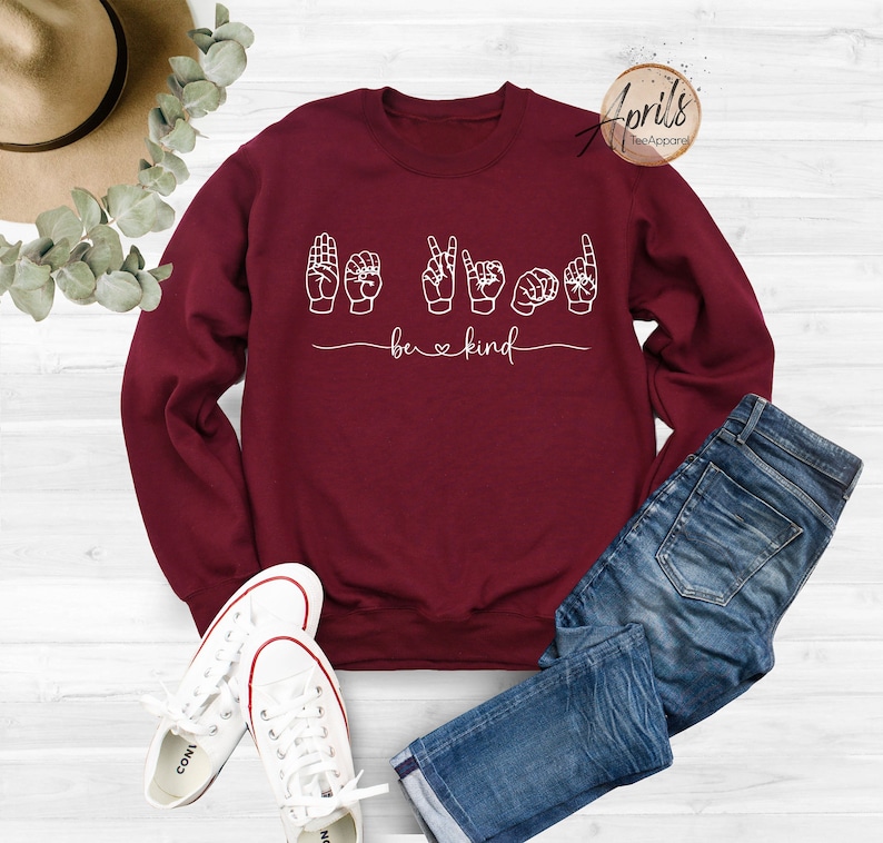 May include: Burgundy sweatshirt with white text that says "be kind" and a sign language interpretation of the phrase.