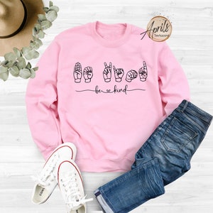 May include: Pink sweatshirt with the words "be kind" in black lettering. The word "be" is in standard lettering and the word "kind" is spelled out using American Sign Language hand gestures.