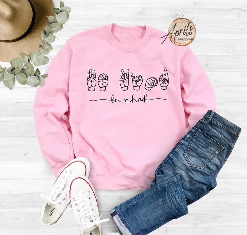 May include: Pink sweatshirt with the words "be kind" in black lettering. The word "be" is in standard lettering and the word "kind" is spelled out using American Sign Language hand gestures.