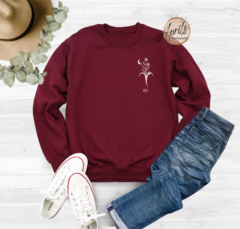 Op de afbeelding: Een bordeauxrood sweatshirt met een witte afbeelding van een bloem en een halve maan met het woord "Aries" eronder. Het sweatshirt ligt op een wit oppervlak met een paar witte sneakers en een blauwe jeans.