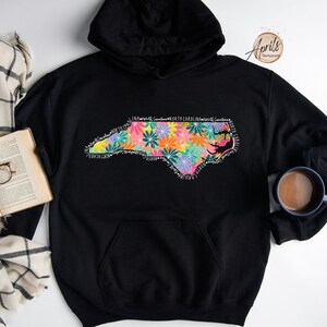 May include: Black hoodie with a floral design of the state of North Carolina. The design includes the words "In Nature's Garden, North Carolina, Heart of Carolina".