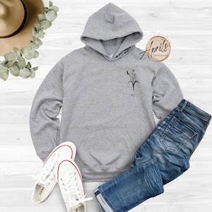 Op de afbeelding: Grijs hoodie met een zwart bloemenontwerp en de tekst "April's Tee Apparel" aan de rechterkant van de capuchon.