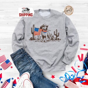 Pu&ograve; includere: Una felpa grigia con una grafica di un cane con un cappello da cowboy e una bandiera americana. Il cane &egrave; in piedi davanti ai cactus. Le parole "Next Day Shipping" sono nell'angolo in alto a sinistra.