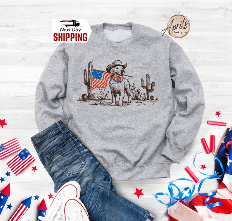 Pu&ograve; includere: Una felpa grigia con una grafica di un cane con un cappello da cowboy e una bandiera americana. Il cane &egrave; in piedi davanti ai cactus. Le parole "Next Day Shipping" sono nell'angolo in alto a sinistra.