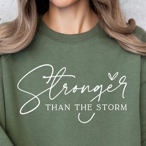 May include: A green sweatshirt with the text "Stronger than the storm" in white cursive lettering with a small heart symbol.