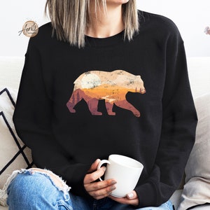 May include: A black sweatshirt with a graphic of a bear silhouette with a sunset and mountains inside the bear. The bear is facing right.