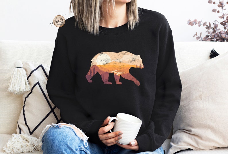 May include: A black sweatshirt with a graphic of a bear silhouette with a sunset and mountains inside the bear. The bear is facing right.