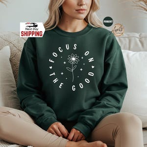 May include: A dark green sweatshirt with the text "FOCUS ON THE GOOD" in a white circular design. The design includes a daisy and small hearts. The sweatshirt is a long-sleeved crew neck.