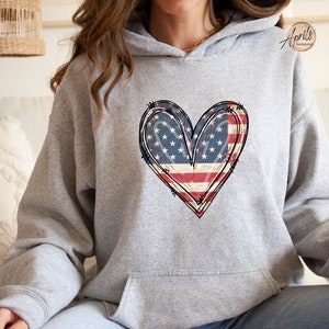 Puede incluir: Una sudadera gris con un dise&ntilde;o de coraz&oacute;n de bandera estadounidense desgastado. El coraz&oacute;n est&aacute; contorneado en negro y tiene un aspecto vintage.