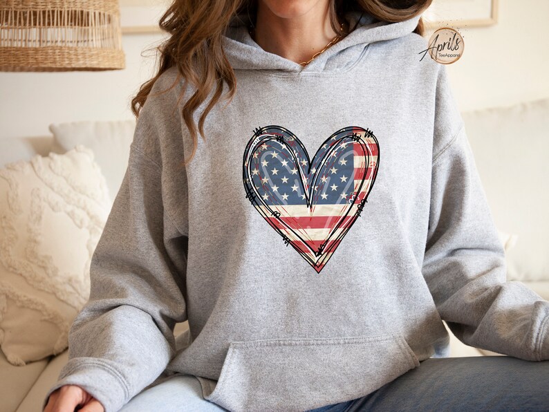 Puede incluir: Una sudadera gris con un dise&ntilde;o de coraz&oacute;n de bandera estadounidense desgastado. El coraz&oacute;n est&aacute; contorneado en negro y tiene un aspecto vintage.