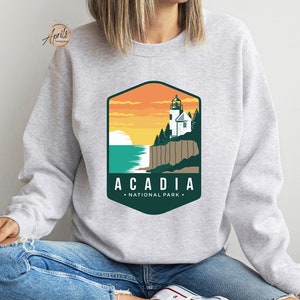 K&ouml;nnte beinhalten: Ein graues Sweatshirt mit einer gr&uuml;nen und orangenen Grafik eines Leuchtturms auf einer Klippe mit Blick auf das Meer. Die Grafik lautet "Acadia National Park".