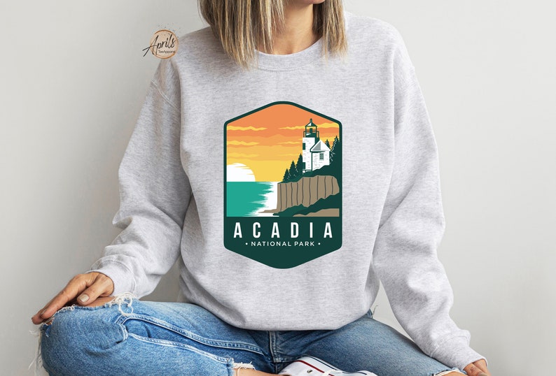 K&ouml;nnte beinhalten: Ein graues Sweatshirt mit einer gr&uuml;nen und orangenen Grafik eines Leuchtturms auf einer Klippe mit Blick auf das Meer. Die Grafik lautet "Acadia National Park".