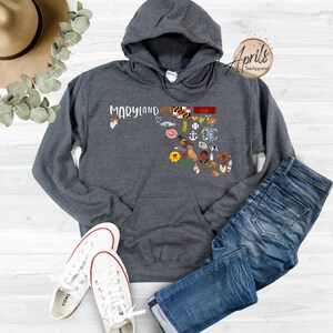 May include: A dark gray hooded sweatshirt with a white outline of the state of Maryland and the word "Maryland" printed on the front. The outline is filled with small icons representing things associated with Maryland, such as the state flag, crabs, and a lighthouse.