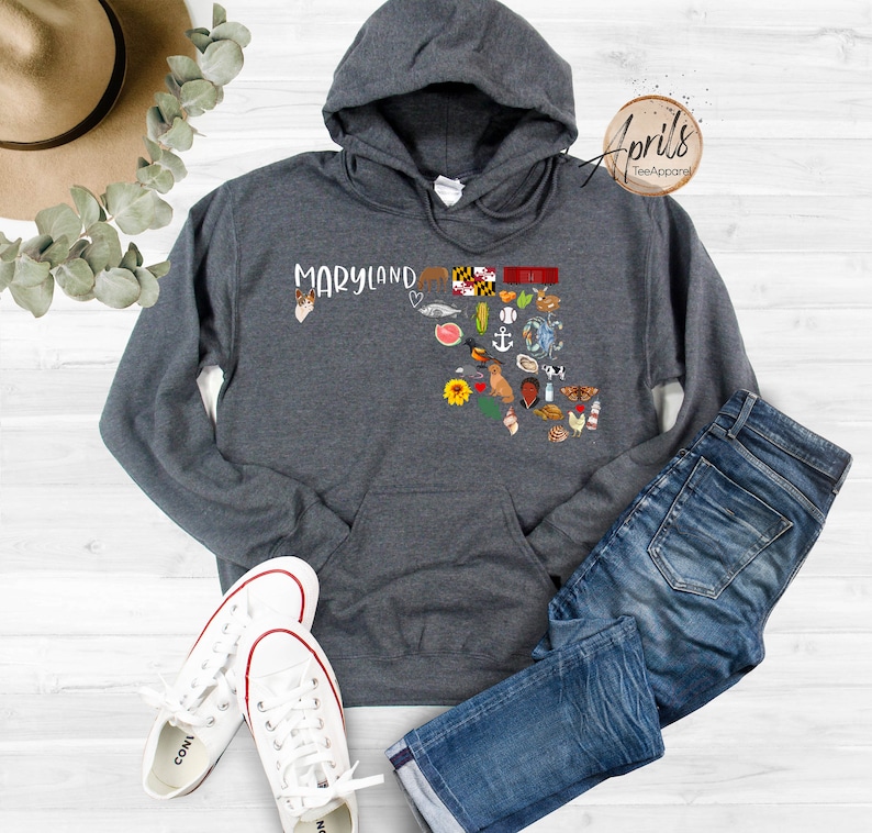 May include: A dark gray hooded sweatshirt with a white outline of the state of Maryland and the word "Maryland" printed on the front. The outline is filled with small icons representing things associated with Maryland, such as the state flag, crabs, and a lighthouse.