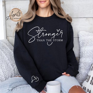 May include: Black sweatshirt with white text that reads "Stronger than the storm" and a small heart. The sweatshirt has a small heart with a cross inside on the sleeve.