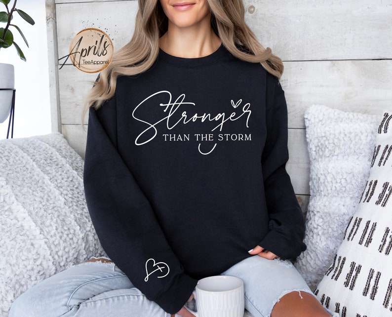 May include: Black sweatshirt with white text that reads "Stronger than the storm" and a small heart. The sweatshirt has a small heart with a cross inside on the sleeve.