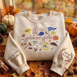 Puede incluir: Una sudadera de cuello redondo color crema con el texto Gilmore Girls en el centro, rodeado de iconos coloridos que incluyen una taza de café, un mazo, una calabaza y letreros de Luke's y Doose's Market. Las mangas presentan el texto where you lead i will follow y I GOT HIT BY A DEER con astas de ciervo.