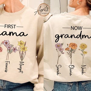 May include: Two beige sweatshirts with floral designs and text. One says "First Mama" with the names James, Francis, and Margot below the flowers. The other says "Now Grandma" with the names Sarah, Olivia, Noah, and Diana below the flowers.