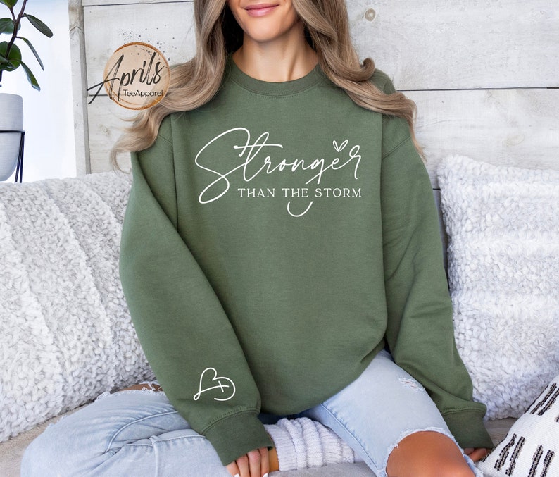 May include: A green sweatshirt with the text "Stronger than the storm" in white lettering. The sweatshirt has a small heart with a cross inside on the left sleeve.