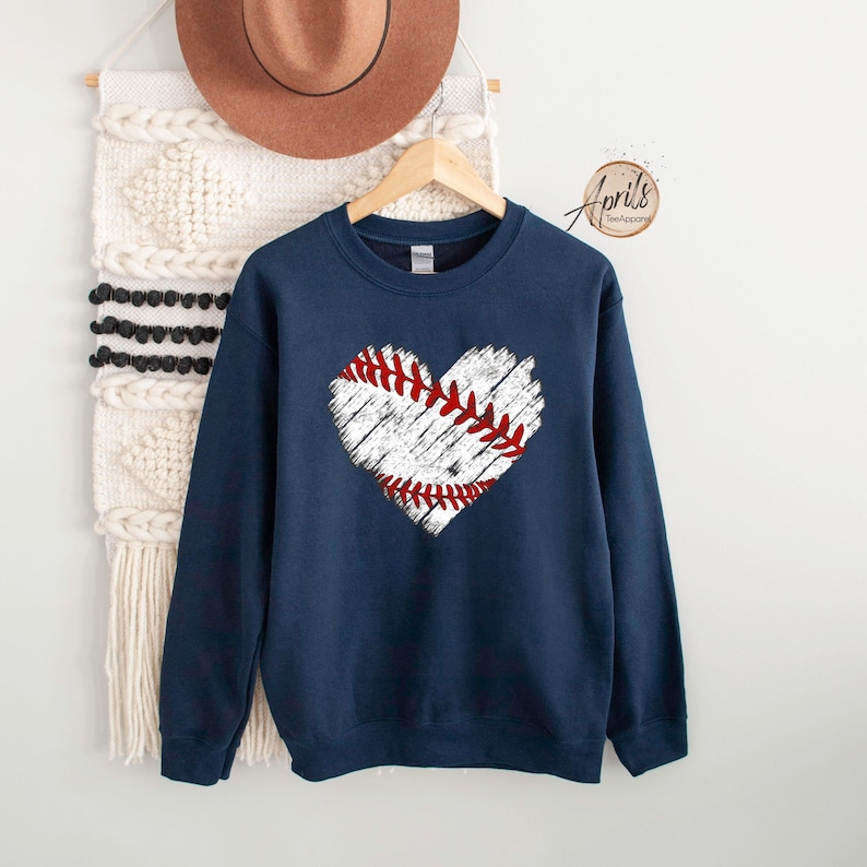 May include: Navy blue sweatshirt with a white distressed heart-shaped baseball graphic. The baseball has red stitching.
