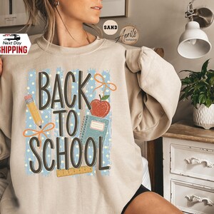May include: A sand-colored crewneck sweatshirt featuring a graphic print with the text BACK TO SCHOOL in dark brown letters. The design includes a yellow pencil, a red apple, a blue spiral notebook, a wooden ruler, and orange ribbon accents.