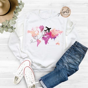 May include: White sweatshirt with a pink and orange watercolor world map design. A black airplane flies over the map with a dashed line leading to two hearts.