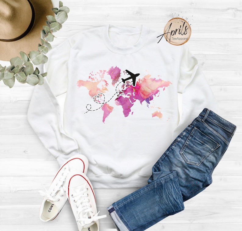 May include: White sweatshirt with a pink and orange watercolor world map design. A black airplane flies over the map with a dashed line leading to two hearts.