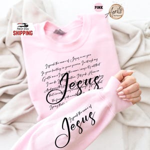 Puede incluir: Sudadera rosa claro con la palabra "Jes&uacute;s" en cursiva. La sudadera tiene un poema impreso en la parte delantera y las palabras "Yo pronuncio el nombre de Jes&uacute;s" en la manga. La sudadera es de color rosa claro.
