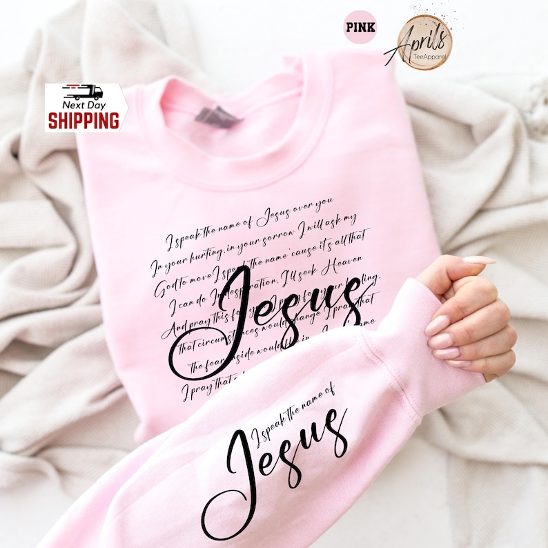 Puede incluir: Sudadera rosa claro con la palabra "Jes&uacute;s" en cursiva. La sudadera tiene un poema impreso en la parte delantera y las palabras "Yo pronuncio el nombre de Jes&uacute;s" en la manga. La sudadera es de color rosa claro.