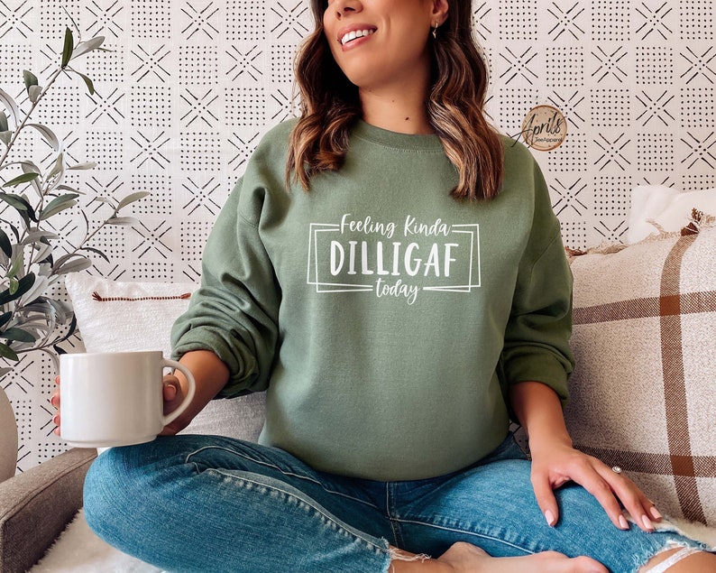May include: A woman wearing a green sweatshirt with the text "Feeling Kinda Dilligaf Today" in white. She is sitting on a couch with a white throw pillow and a brown plaid throw pillow.
