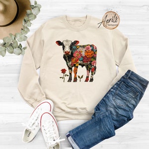 May include: A beige sweatshirt with a floral cow design. The cow is black and white with colorful flowers covering its body. The sweatshirt is shown on a white surface with a pair of blue jeans and white sneakers.
