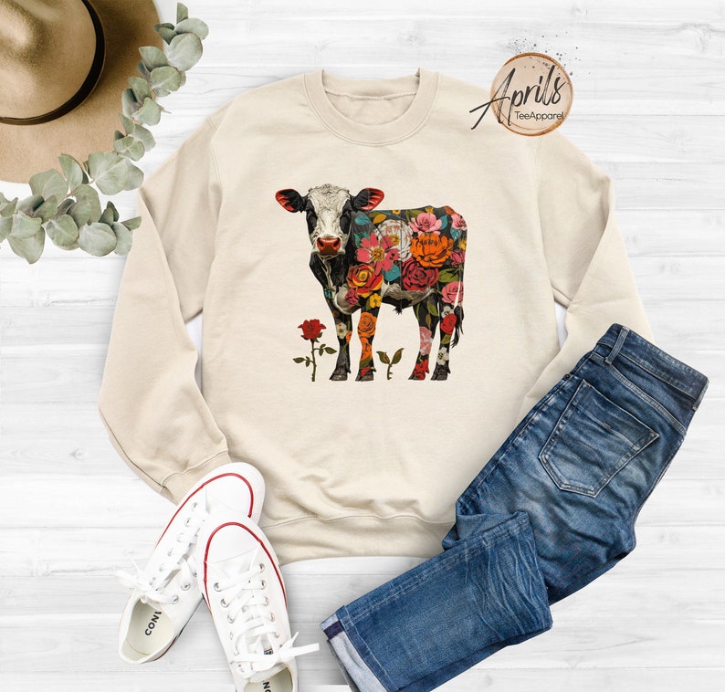 May include: A beige sweatshirt with a floral cow design. The cow is black and white with colorful flowers covering its body. The sweatshirt is shown on a white surface with a pair of blue jeans and white sneakers.