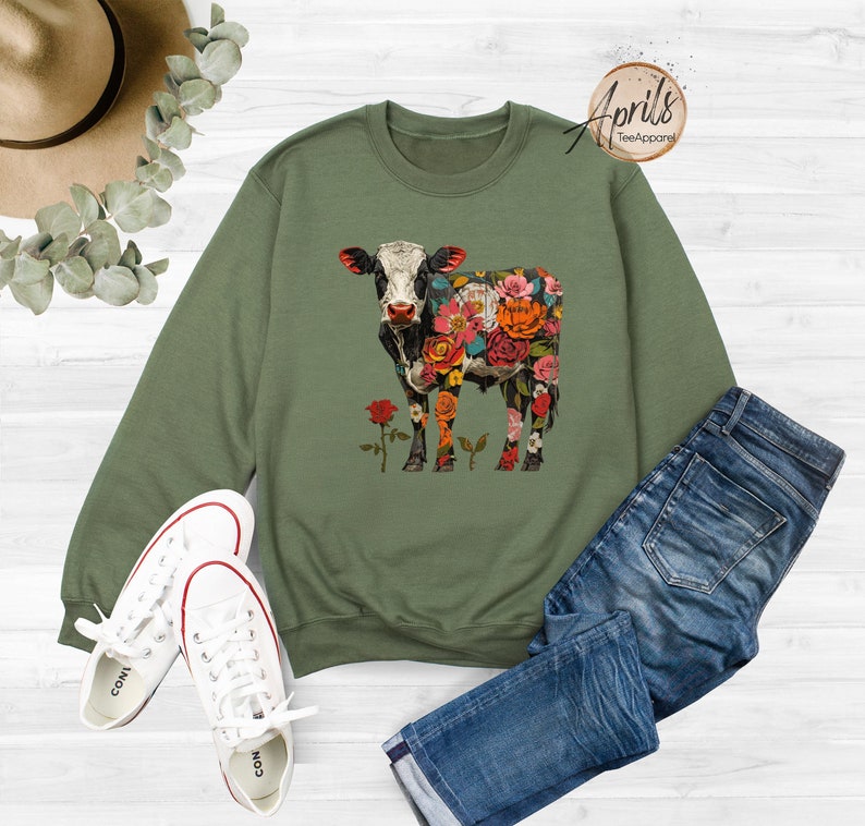 May include: A green sweatshirt with a floral cow design. The cow is black and white with colorful flowers covering its body. The sweatshirt is laid out on a white surface with a pair of white sneakers and blue jeans.