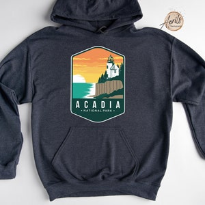 K&ouml;nnte beinhalten: Ein dunkelblauer Kapuzenpullover mit einem grafischen Design eines Leuchtturms und dem Text "Acadia National Park".