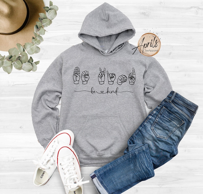 May include: Gray hooded sweatshirt with the words "be kind" in black lettering and a heart symbol. The word "be" is signed in American Sign Language.