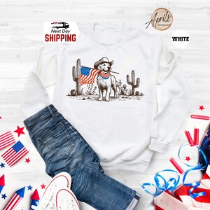 Pu&ograve; includere: Felpa bianca con grafica di un cane con cappello da cowboy e bandiera americana. Il cane &egrave; davanti ai cactus. Le parole "Next Day Shipping" sono stampate in alto a sinistra.