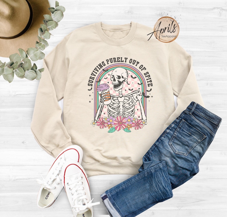 May include: A beige sweatshirt featuring a graphic of a skeleton holding a coffee cup. The graphic reads "Surviving purely out of spite" with a rainbow and bats surrounding the skeleton.