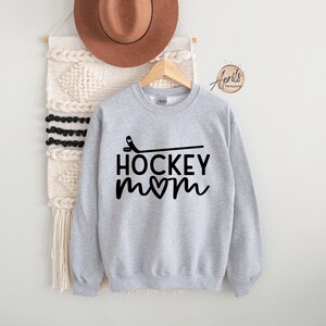 May include: Gray sweatshirt with black text that reads "Hockey Mom" with a heart in the "o". A hockey stick is drawn above the text.