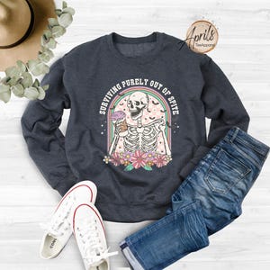 May include: A dark blue sweatshirt featuring a graphic of a skeleton holding a coffee cup. The graphic reads "Surviving purely out of spite".