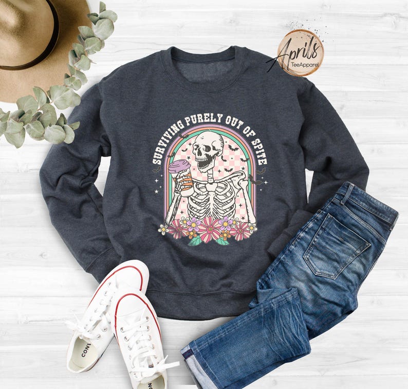 May include: A dark blue sweatshirt featuring a graphic of a skeleton holding a coffee cup. The graphic reads "Surviving purely out of spite".