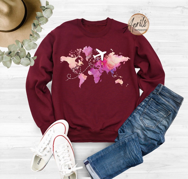 May include: Burgundy sweatshirt with a watercolor world map design and a white airplane flying over it. The map has pink, purple, and blue hues. The text "Aprils Tee Apparel" is in the top right corner.