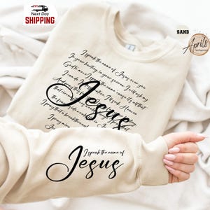 Puede incluir: Sudadera de color arena con la palabra "Jes&uacute;s" en una fuente grande y cursiva. La sudadera presenta un poema sobre Jes&uacute;s en escritura negra. La manga muestra el texto "Yo pronuncio el nombre de Jes&uacute;s."