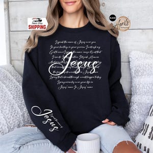 Puede incluir: Sudadera negra con la palabra "Jes&uacute;s" en cursiva. La sudadera tiene un poema impreso en la parte delantera y las palabras "Hablo el nombre de Jes&uacute;s" en la manga. La sudadera la lleva una persona.