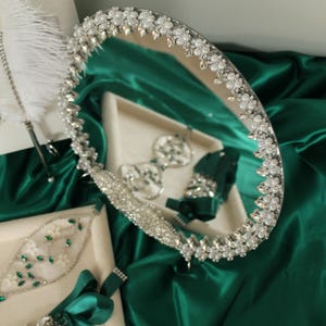 May include: A round mirror with a decorative border of white pearls and clear crystals, accompanied by a white feather pen and several green satin ribbon accessories adorned with matching crystals and pearls on cream-colored display trays.