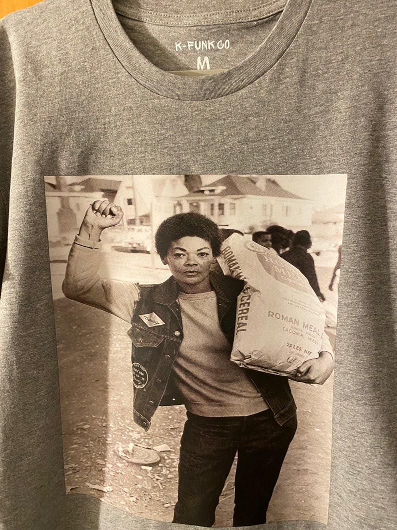 May include: A black and white photograph of a woman holding a bag of Roman Meal cereal. The woman is wearing a denim jacket and jeans. The bag of cereal has the text "Roman Meal" and "25 lbs. Net" printed on it.