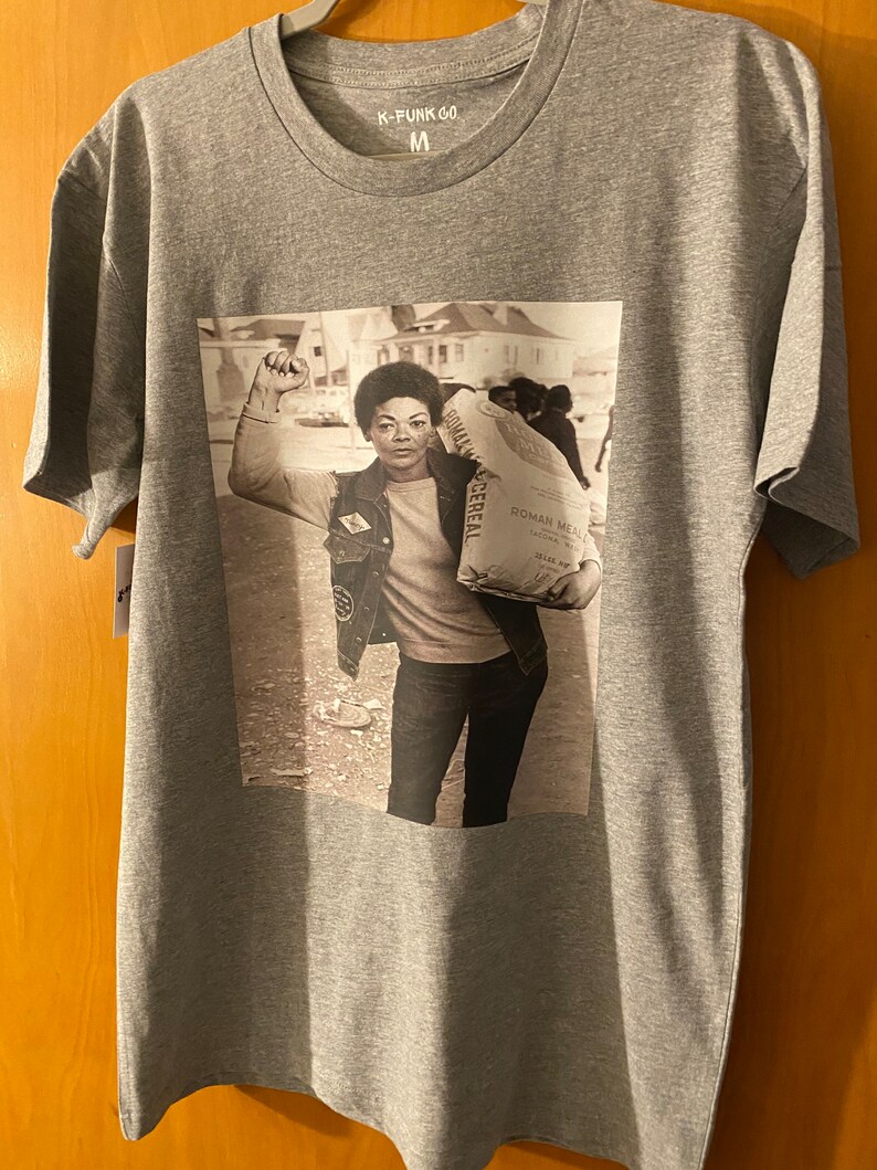 May include: A gray t-shirt with a black and white photo of a woman holding a bag of flour. The bag of flour has the text "Roman Meal" printed on it. The t-shirt has the text "K-Funk Co" printed on the tag.