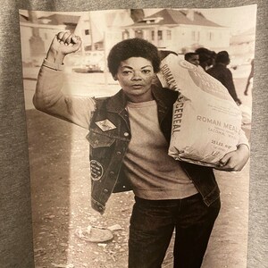 May include: A black and white photograph of a woman raising her fist in the air. She is wearing a denim jacket with a patch that says "Timothy" and is carrying a bag of Roman Meal Cereal. The bag is labeled "Roman Meal Cereal, Enriched, 25 lbs. Net, Tacoma, Wash."
